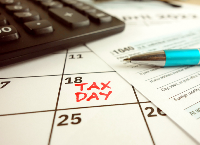 Close up of a desk calendar with TAX DAY written in red on the 18th day. A tax form, pen & computer keyboard lays on top.